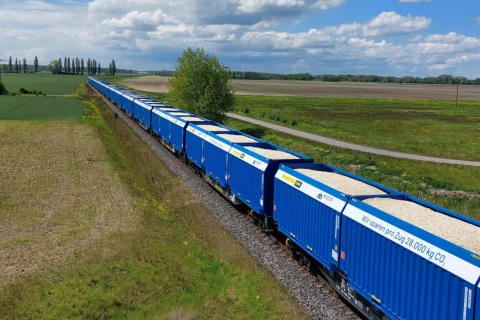 Ein langer blauer Güterzug voller hellem Schüttgut fährt durch grüne Felder. Waggons bewerben 28.000 kg CO₂-Einsparung pro Zug.