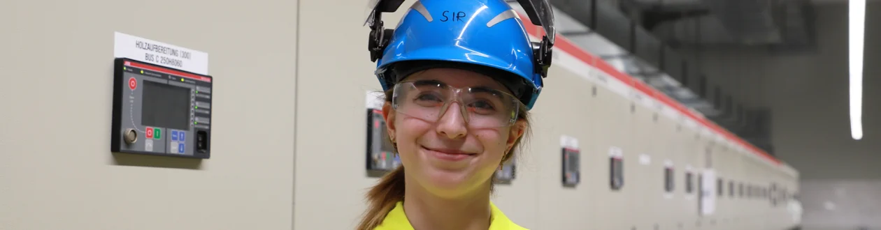 Lächelnde junge Frau in gelber PSA, blauem Helm und Schutzbrille in einer Industrieanlage mit Schaltschränken.
