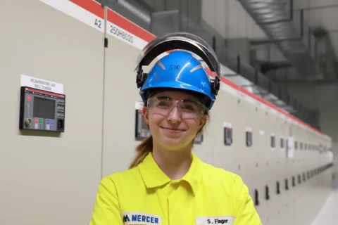 Lächelnde junge Frau in gelber PSA, blauem Helm und Schutzbrille in einer Industrieanlage mit Schaltschränken.