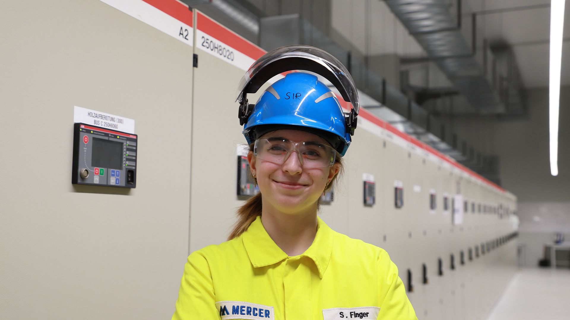 Lächelnde junge Frau in gelber PSA, blauem Helm und Schutzbrille in einer Industrieanlage mit Schaltschränken.