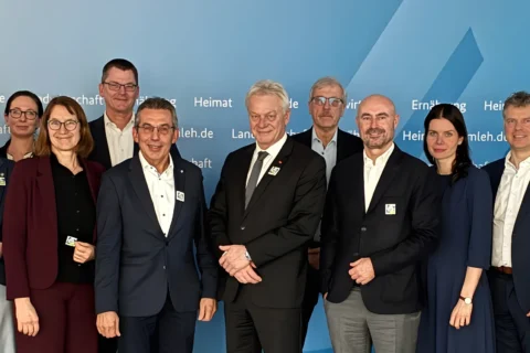 17 Personen (13 Männer, 4 Frauen) in Businesskleidung lächeln vor blauer Wand mit Text. Viele tragen grüne Abzeichen.