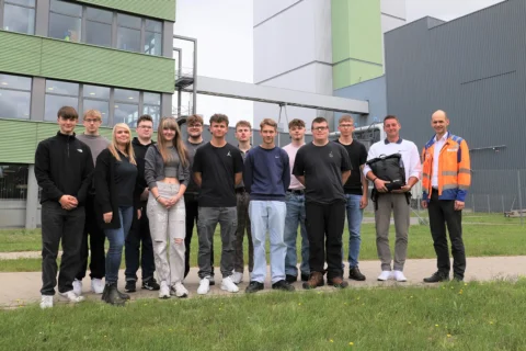 15 Personen (Jugendliche & 2 Erwachsene) stehen vor Industriegebäuden. Einer trägt Warnweste. Schulung oder Exkursion.