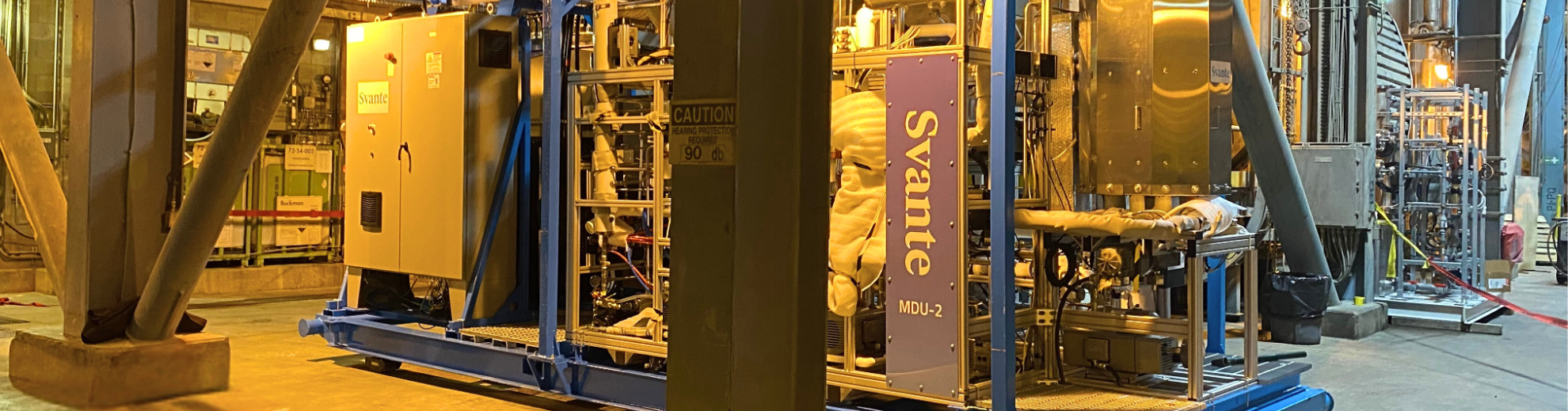 Komplexes Svante MDU-2 Prozessmodul auf blauem Rahmen in Industriehalle. Massiver Träger, Rohre, Kräne unter warmem gelben Licht.