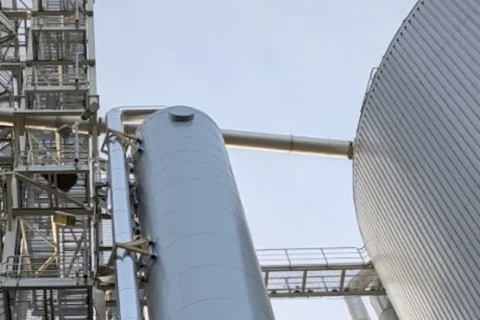 Industrieanlage: Silberne Tanks, Zylinder, Rohre und Gerüste dominieren das Bild, dazu ein großes, geripptes Gebäude.