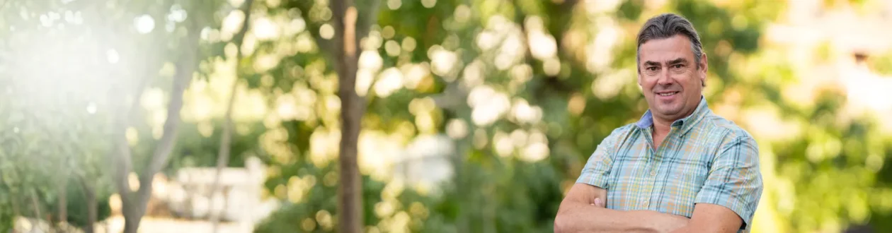 Porträt eines lächelnden Mannes mit grauem Haar, kariertem Hemd und verschränkten Armen vor unscharfem Grün und Sonnenlicht.