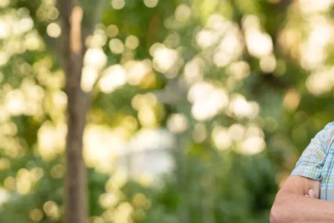 Porträt eines lächelnden Mannes mit grauem Haar, kariertem Hemd und verschränkten Armen vor unscharfem Grün und Sonnenlicht.