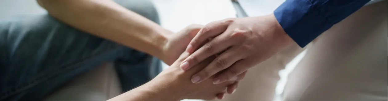 Nahaufnahme: Eine Hand im blauen Hemd hält tröstend die Hand einer Person in dunkler Kleidung. Symbolisiert Unterstützung.
