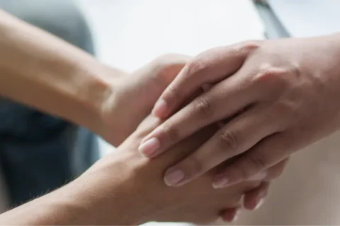 Nahaufnahme: Eine Hand im blauen Hemd hält tröstend die Hand einer Person in dunkler Kleidung. Symbolisiert Unterstützung.