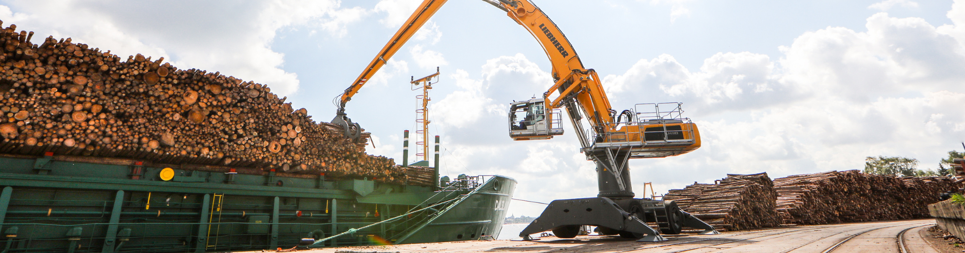 Ein orangefarbener Liebherr-Kran lädt Holzstämme von einem großen Stapel auf ein grünes Frachtschiff in einem Hafen unter bewölktem Himmel.